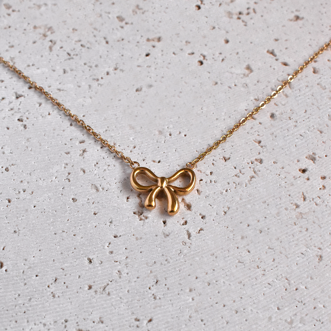 Stainless Steel Bowtiful Necklace. This lovely and trendy piece features a delicate bow ribbon design, plated with stunning 18k gold. Add a touch of sophistication to any outfit with this statement accessory.
